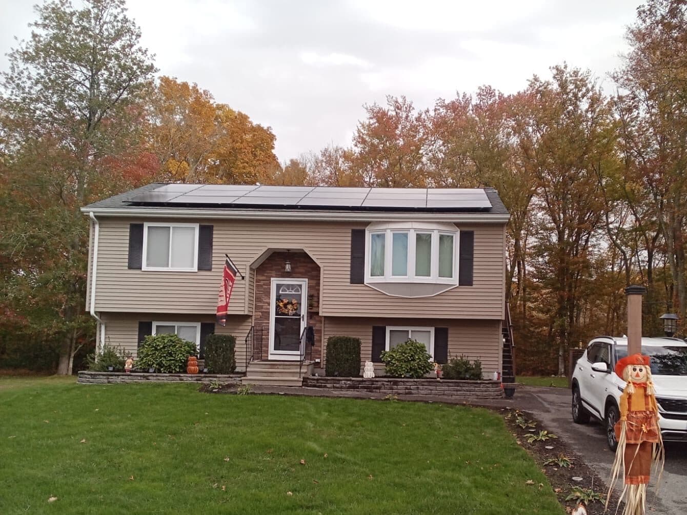 A split-level home with tan siding and black shutters, featuring rooftop solar panels. The house has a small front porch decorated with seasonal items, while the yard displays fall decorations, including a scarecrow near the driveway.