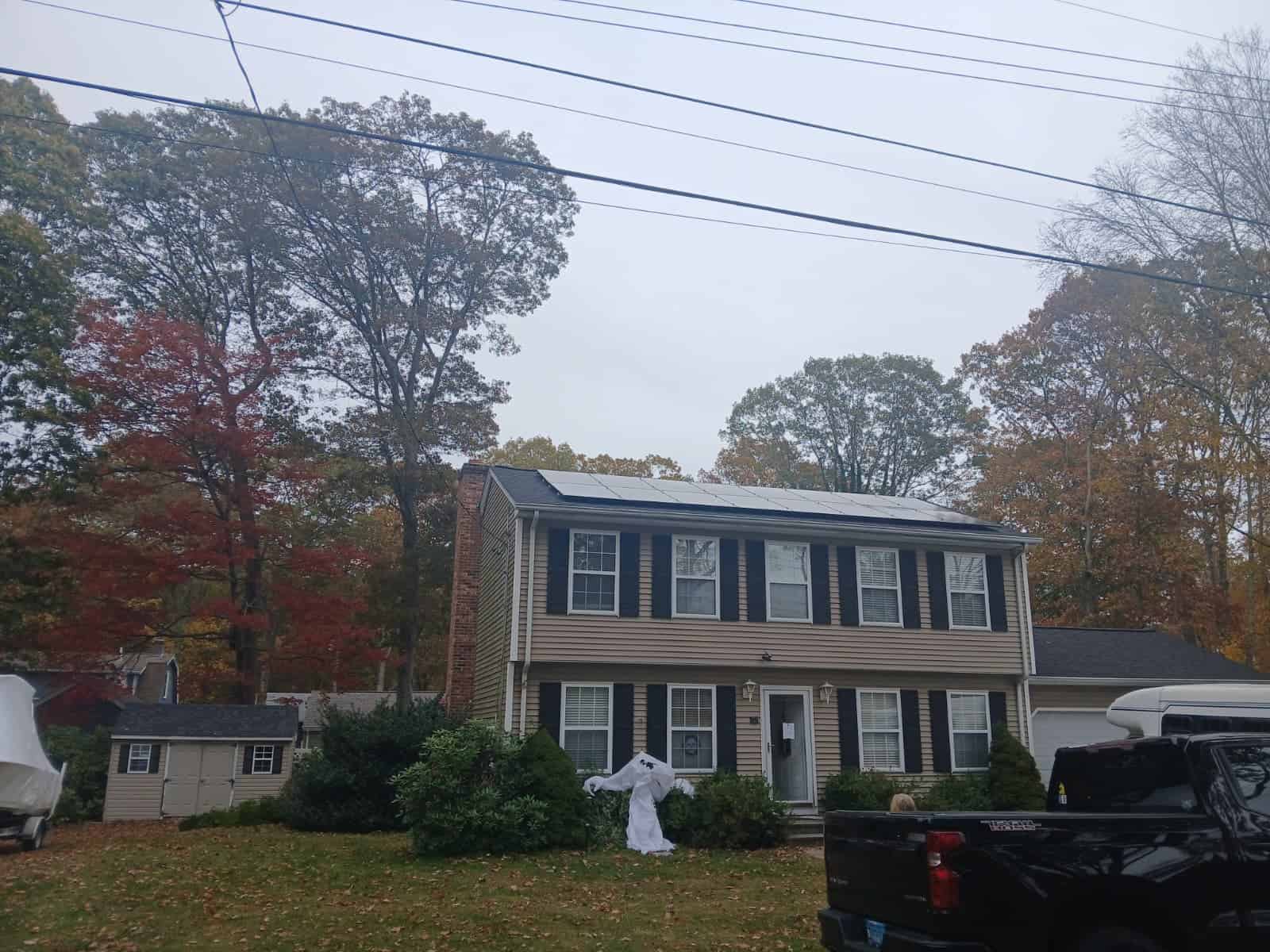 A two-story colonial home with tan siding and black shutters, featuring a solar panel array installed across the front roof, surrounded by autumn trees.