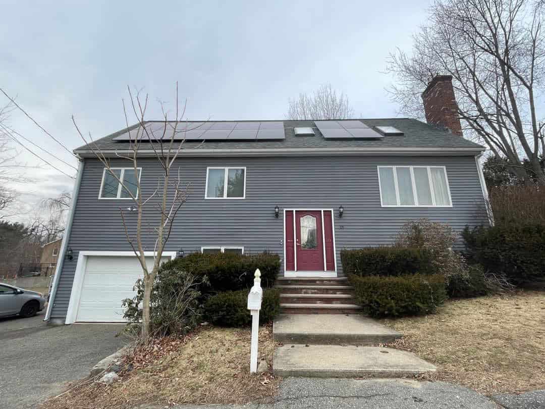 Solar panels installed on the roof of a gray, two-story house with a red front door, set on a slightly sloped yard with bushes and a tree, capturing renewable energy from sunlight.