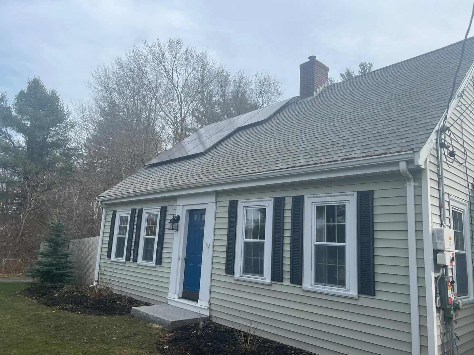 Roof-mounted solar panels installed on a suburban home with beige siding, black shutters, and a blue front door, surrounded by a landscaped lawn and trees, showcasing residential solar energy solutions.