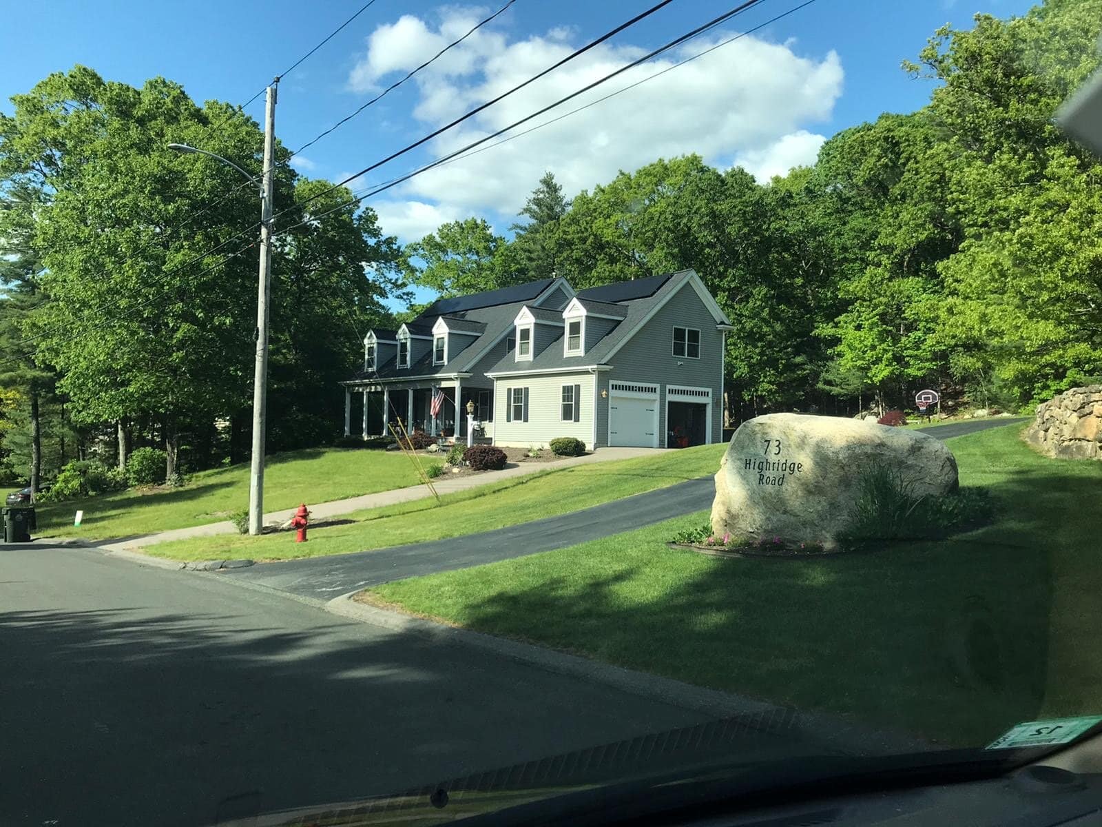 Large traditional two-story gray house with white trim and black roof, surrounded by lush green trees, located at 73 Highridge Road with a prominent rock displaying the address, under a blue sky with scattered clouds.