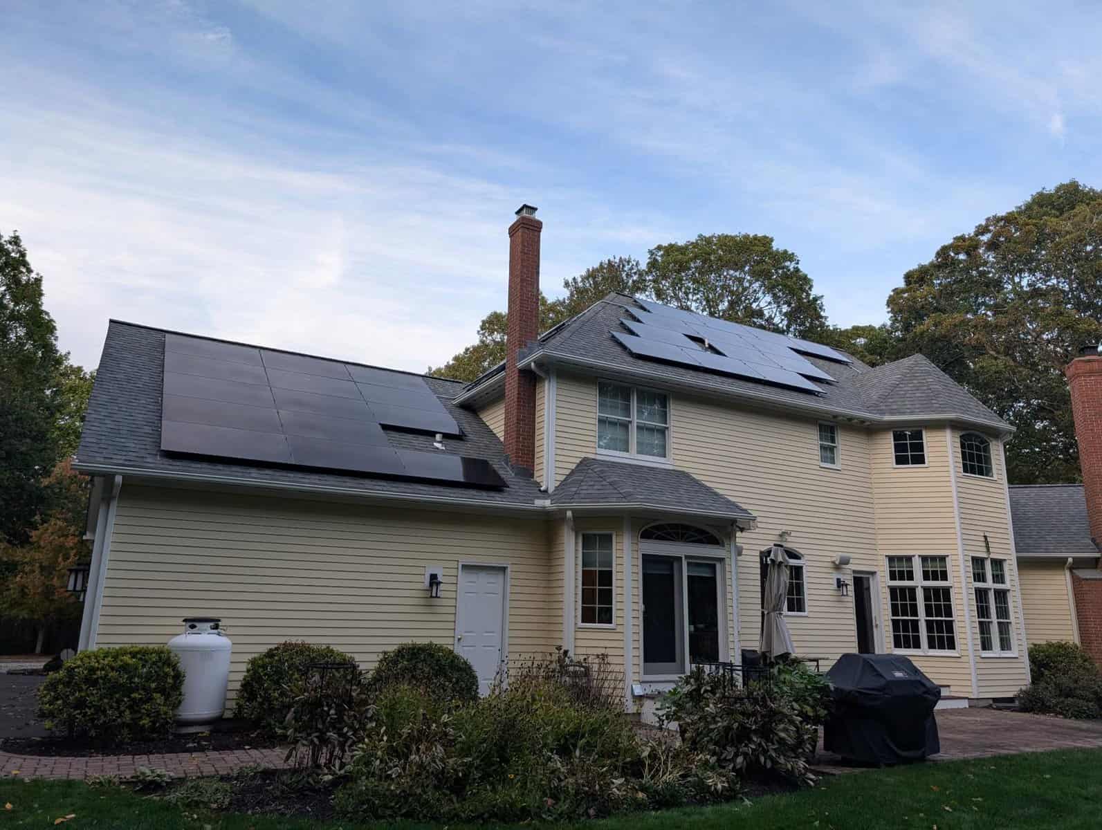 Large two-story New England home with beige siding and multiple roof-mounted solar panel arrays, highlighting a professional residential solar installation designed to reduce electric bills, improve home energy efficiency, and maximize long-term solar savings.
