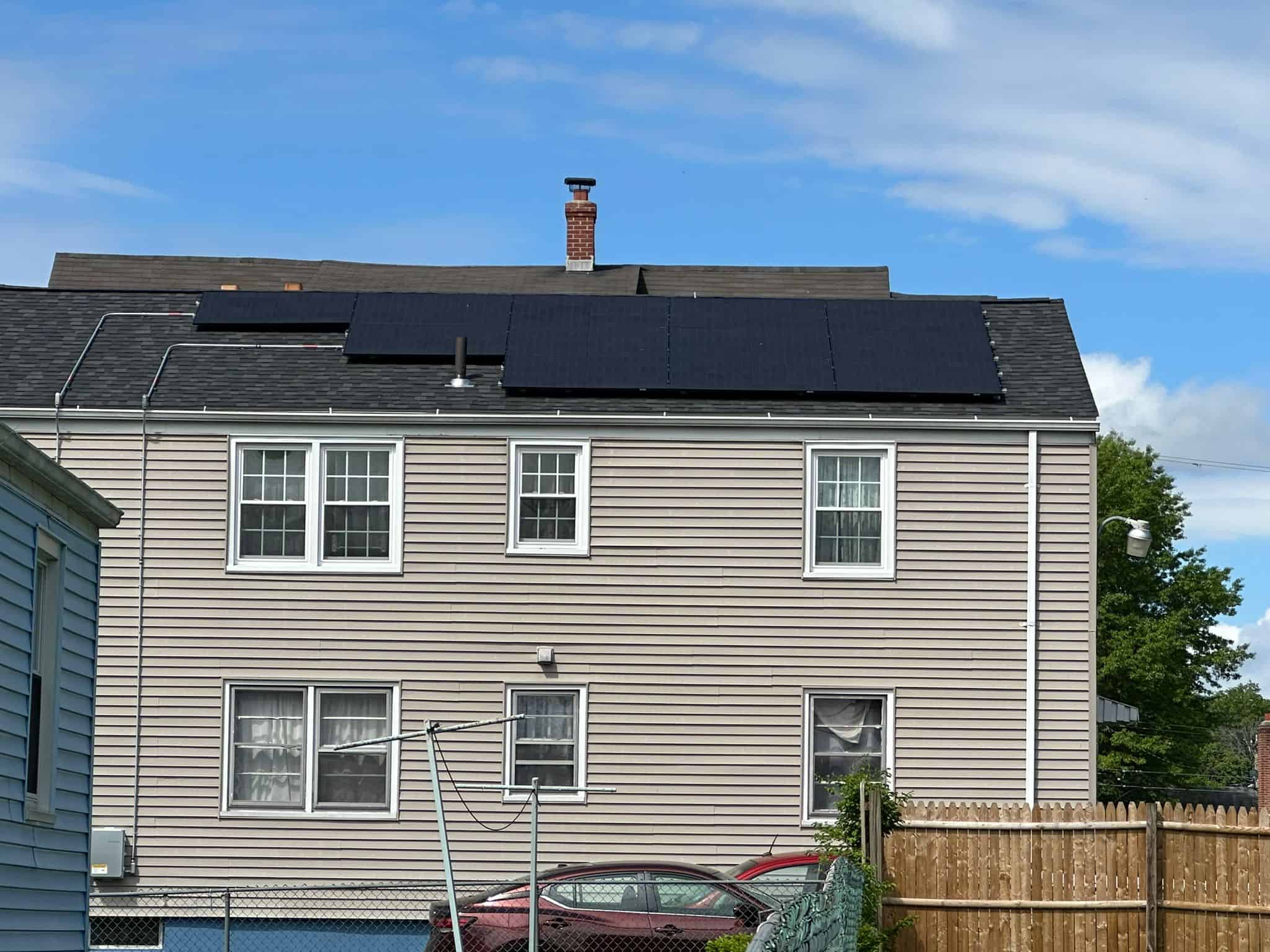 Photovoltaic solar panels installed on the roof of a beige residential house, generating clean energy and reducing electricity bills. The house features multiple windows, a chimney, and a clear blue sky background.