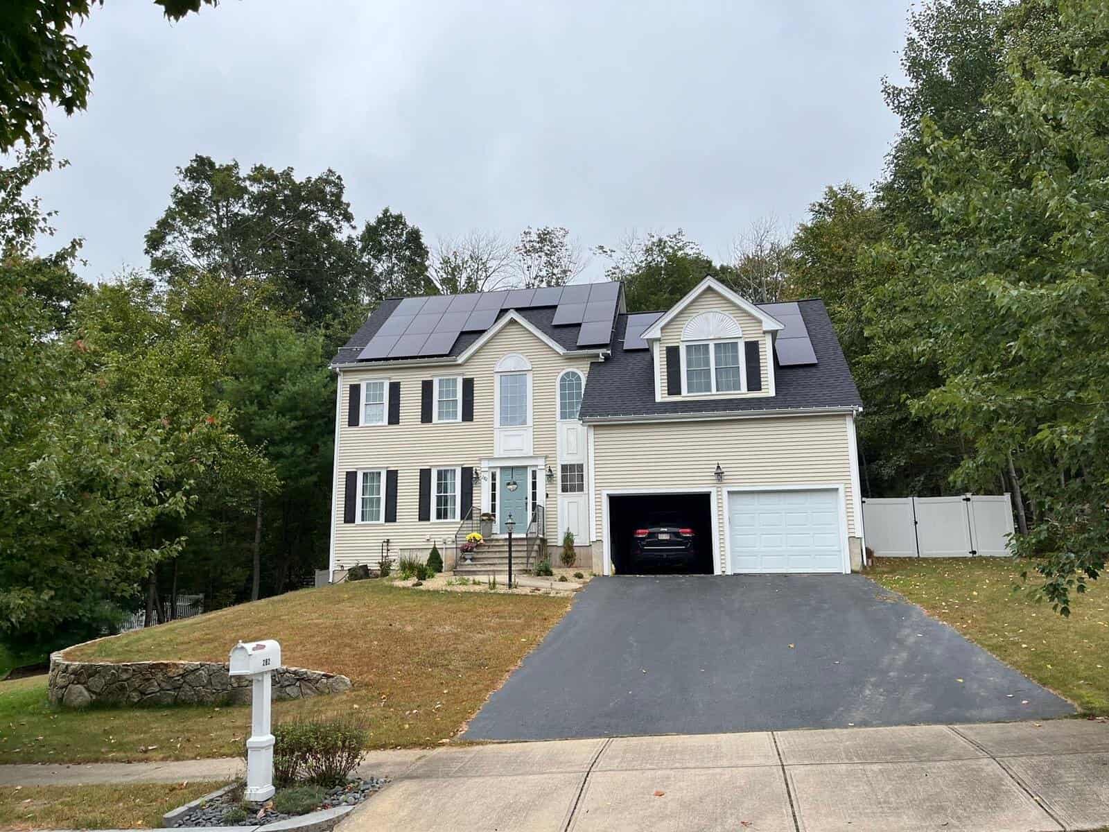 Beige two-story colonial-style home with black shutters and a light blue front door, featuring multiple rooftop solar panels installed across the front-facing rooflines.