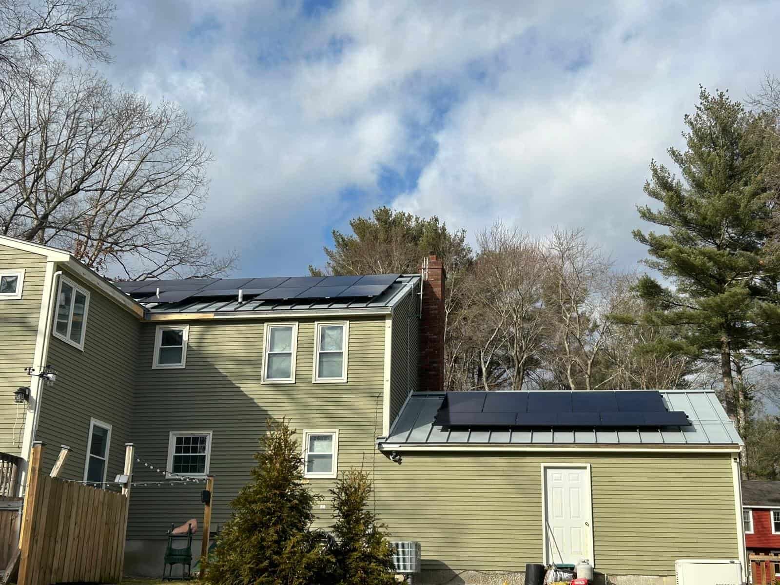 Two-story light green home with both the main roof and a side addition fitted with new solar panels, surrounded by tall trees and a partly cloudy sky.
