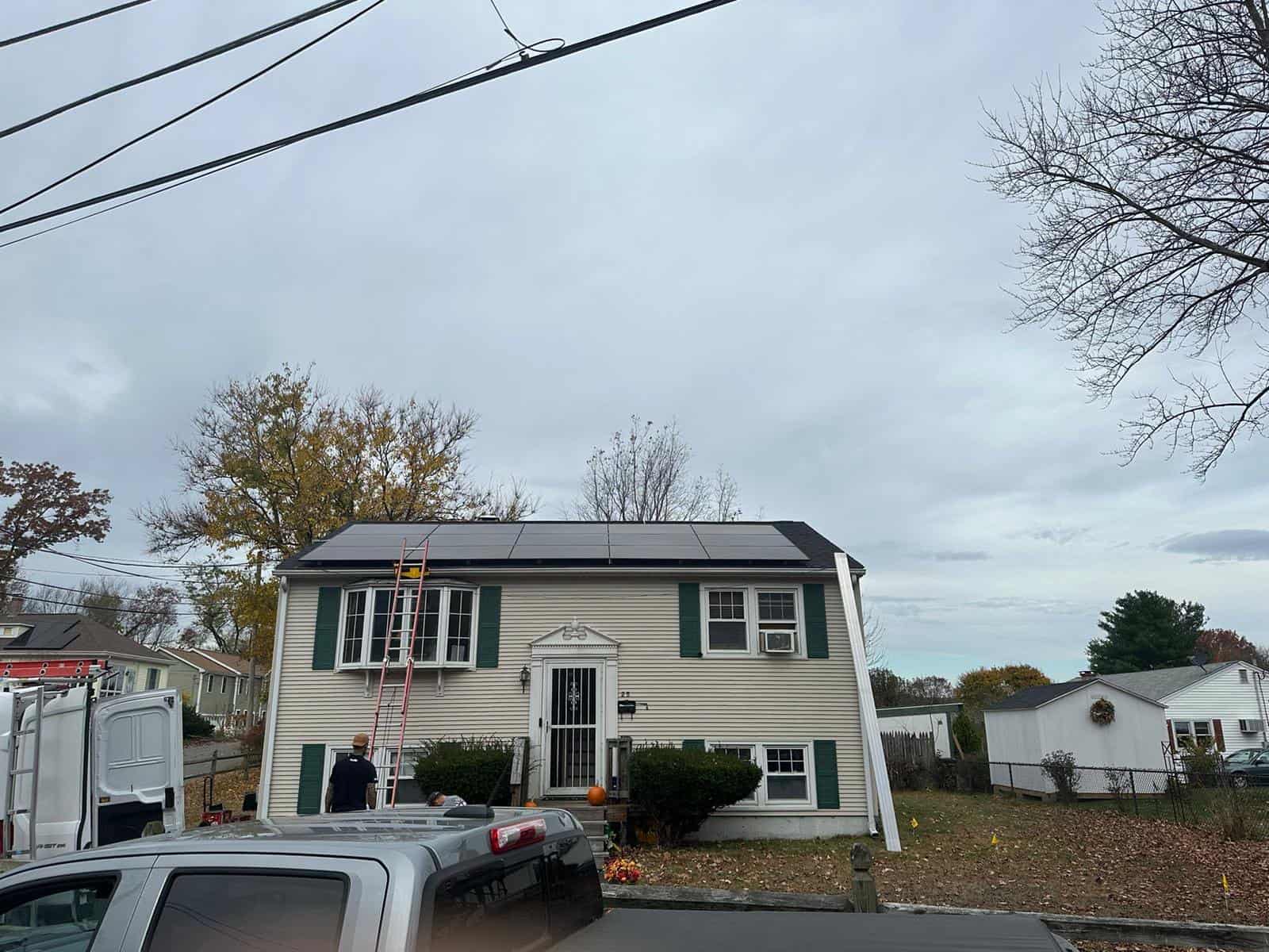 A two-story home with cream siding and green shutters featuring newly installed rooftop solar panels, with installation equipment and vehicles visible in the driveway.