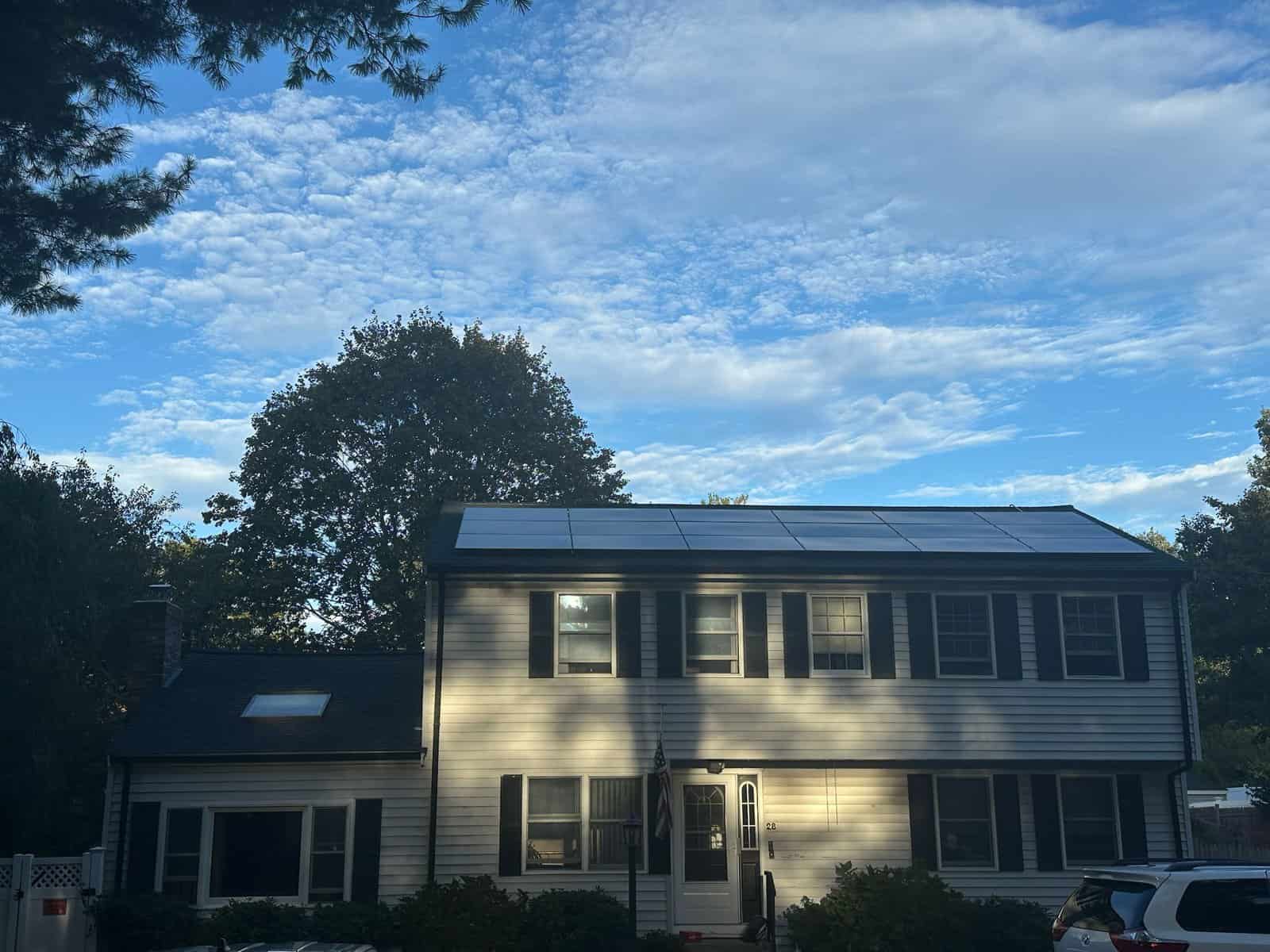 Two-story suburban home with black solar panels installed along the upper roofline, viewed from the front under a blue sky with scattered clouds, showing a residential solar installation integrated into an existing roof.