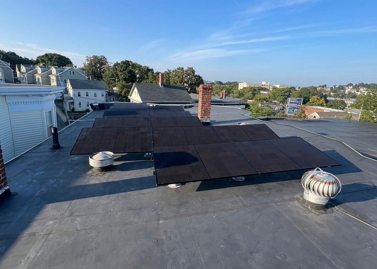 Solar panels installed on a flat rooftop, positioned around chimneys and vents, with a neighborhood of homes visible in the background.