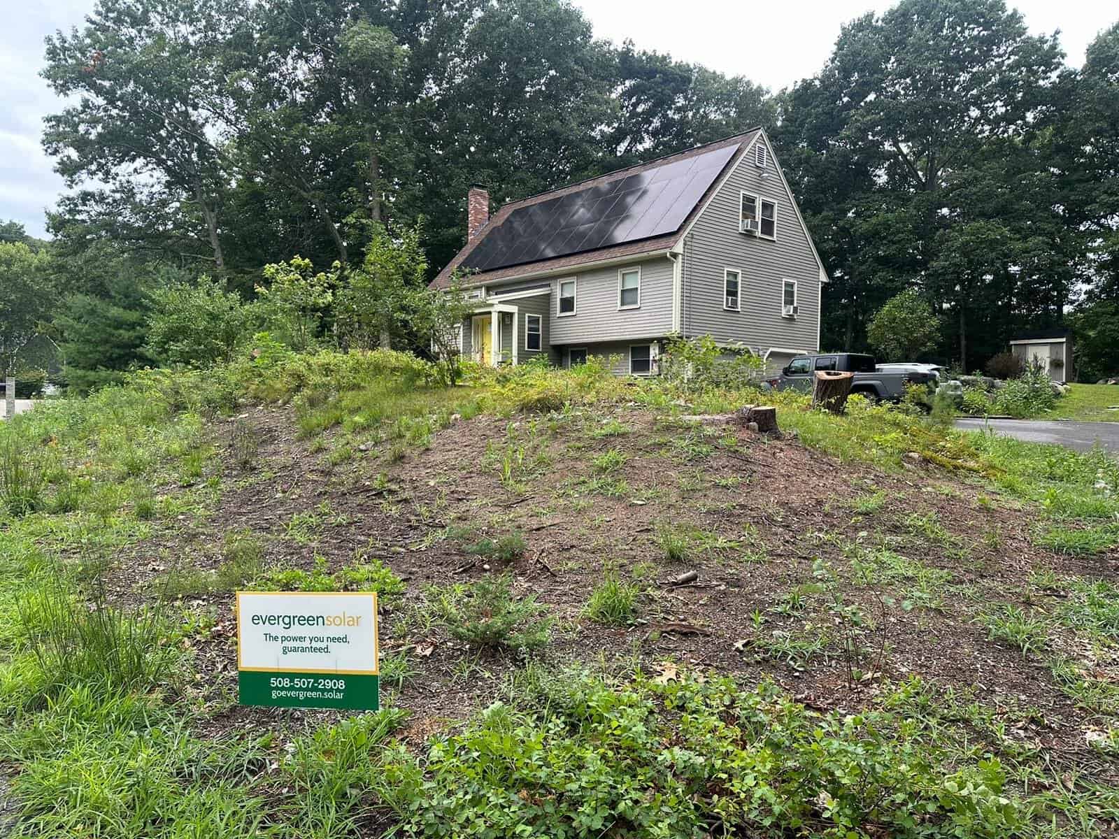 Solar panels cover the steep roof of this gray two-story home, paired with an Evergreen Solar sign displayed in the front yard.