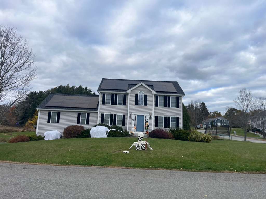 Front exterior view of a two-story New England home with roof-mounted solar panels installed on asphalt shingles, showcasing a residential solar energy system designed to lower electric bills, increase solar savings, and provide long-term energy efficiency for homeowners despite cloudy weather conditions.