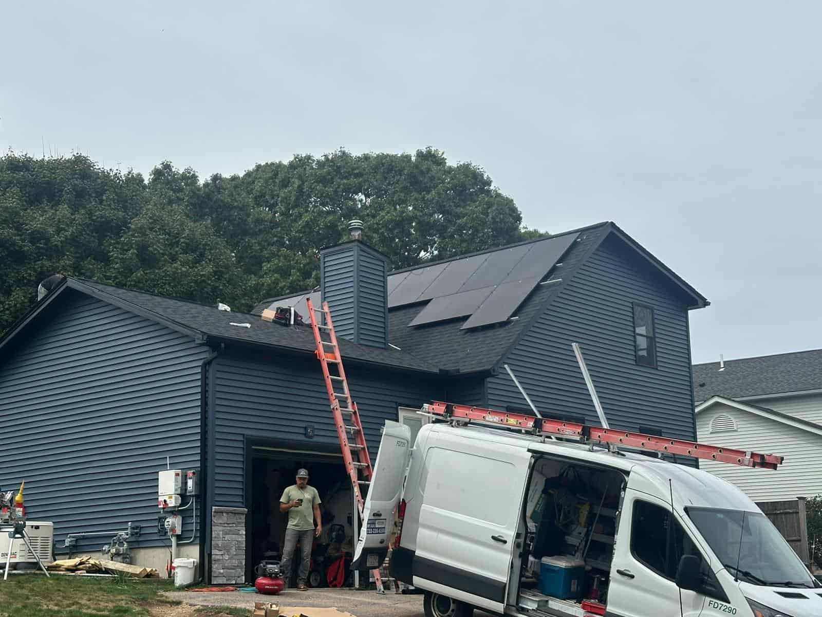 New England home with white siding and a dark shingle roof, featuring solar panels installed on both the main roof and garage roof, overlooking a backyard patio area.