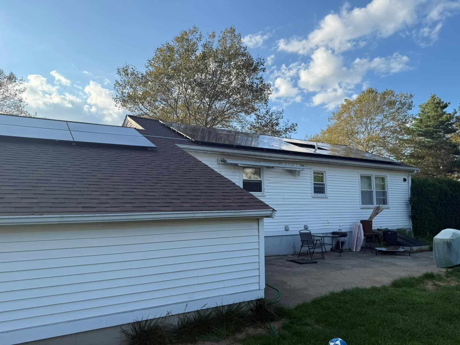 New England home with white siding and a dark shingle roof, featuring solar panels installed on both the main roof and garage roof, overlooking a backyard patio area.