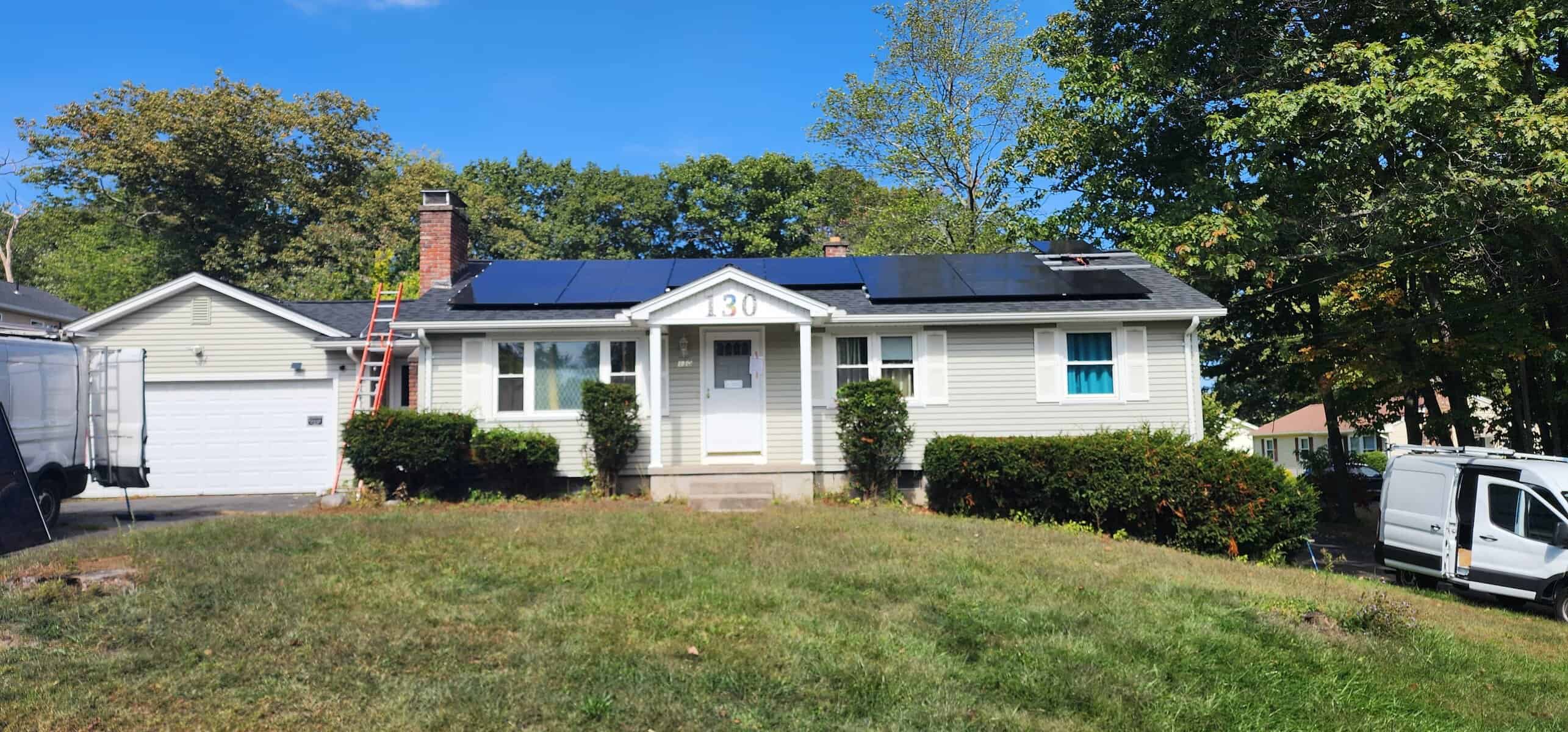 New England ranch-style home with freshly installed solar panels across the roof, work vans and ladders on-site showing the installation in progress under a clear blue sky.