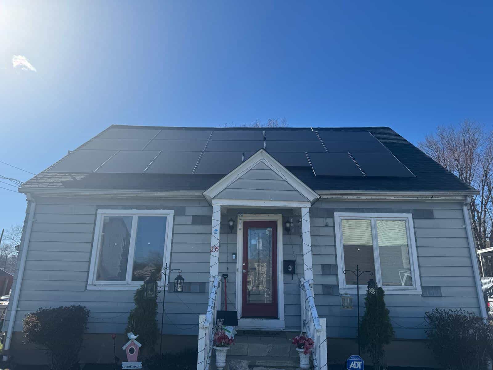 Solar-powered residential house with a dark rooftop solar panel system installed on a clear, sunny day, featuring white exterior, front porch, and well-maintained garden; part of sustainable energy solutions.