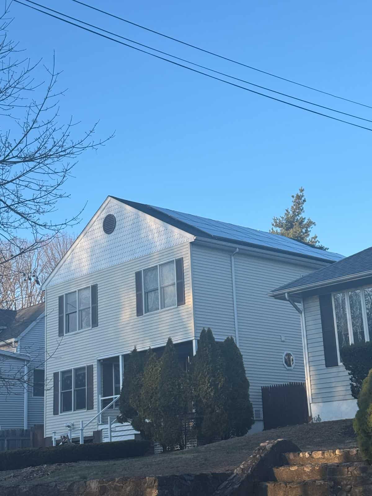 Solar house with roof-mounted photovoltaic panels, white siding, and black shutters under a clear blue sky, surrounded by trees and a landscaped yard, showcasing residential solar energy installation for sustainable power.