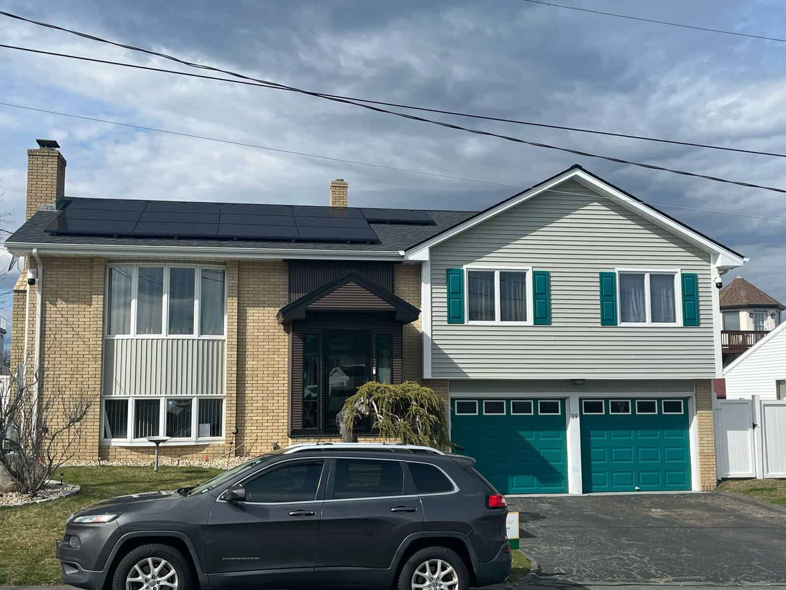 Photovoltaic solar panels installed on the roof of a suburban home, providing clean and renewable energy to reduce electricity costs and environmental impact. The house features a brick and siding exterior with teal-colored shutters.