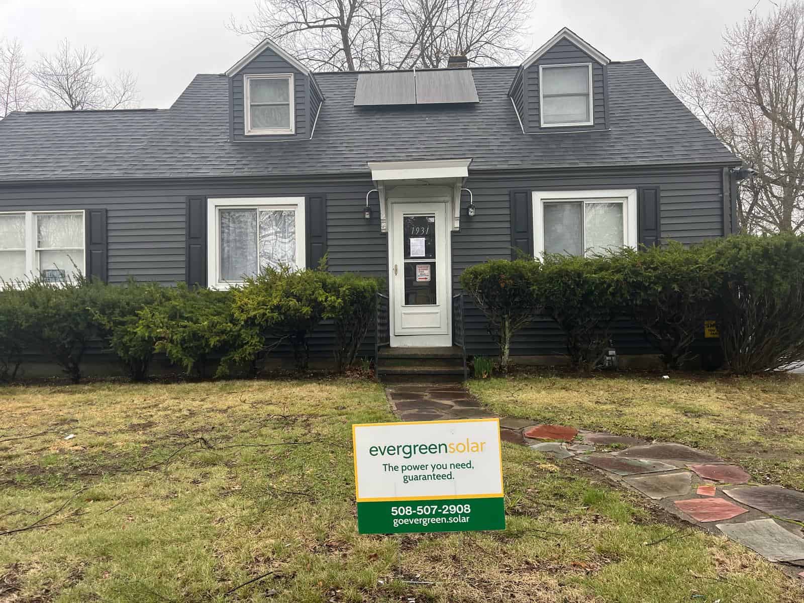 Photovoltaic solar panels installed on the roof of a dark gray house with front garden bushes, small steps leading to the front door, and a company sign promoting Evergreen Solar solar energy solutions.