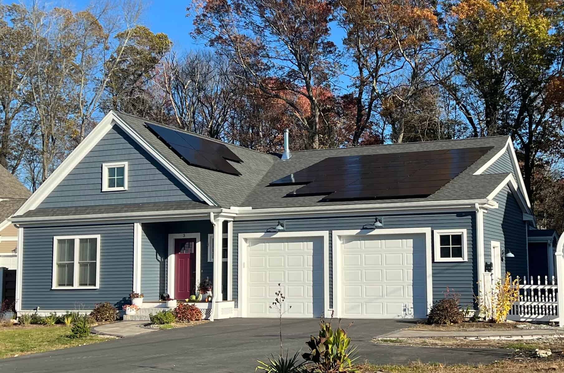 Front exterior of a blue two-story home with an attached two-car garage and a professionally installed rooftop solar panel system. The residential solar panels are flush-mounted on an asphalt shingle roof, blending cleanly with the home’s design and showcasing a modern solar installation surrounded by fall trees.