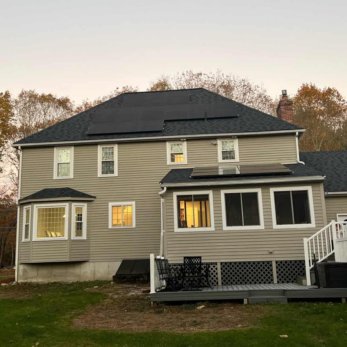 Rear view of a two-story New England home with a dark shingle roof and a flush-mounted solar panel array spanning the upper roof plane. The clean panel layout blends with the roofline while capturing optimal sunlight for efficient residential solar energy production.