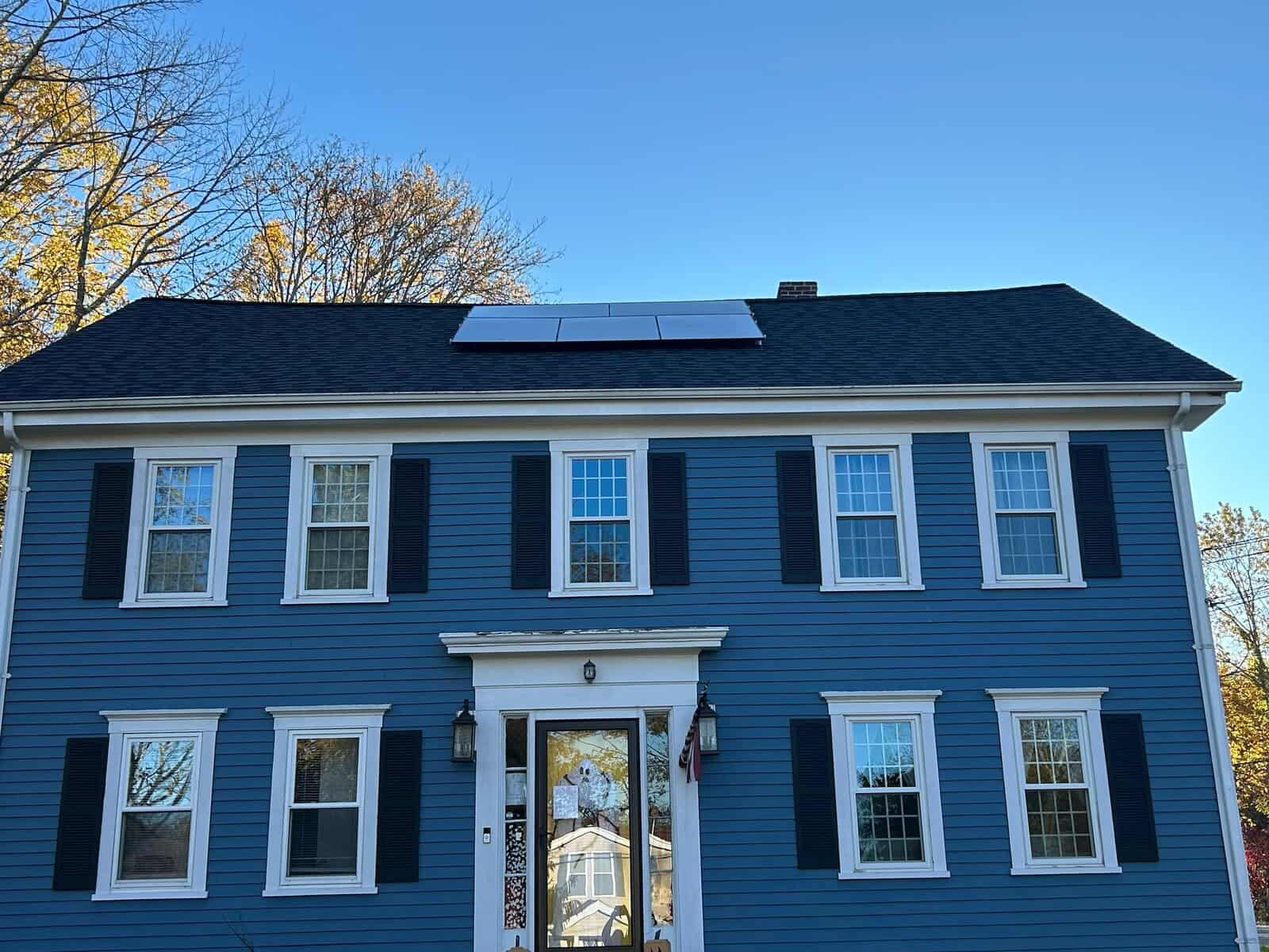 Front-facing view of a blue colonial home with white trim and a low-profile rooftop solar array installed near the roof ridge. The panel layout follows the roofline cleanly, maintaining curb appeal while maximizing sunlight exposure for efficient residential solar energy production.