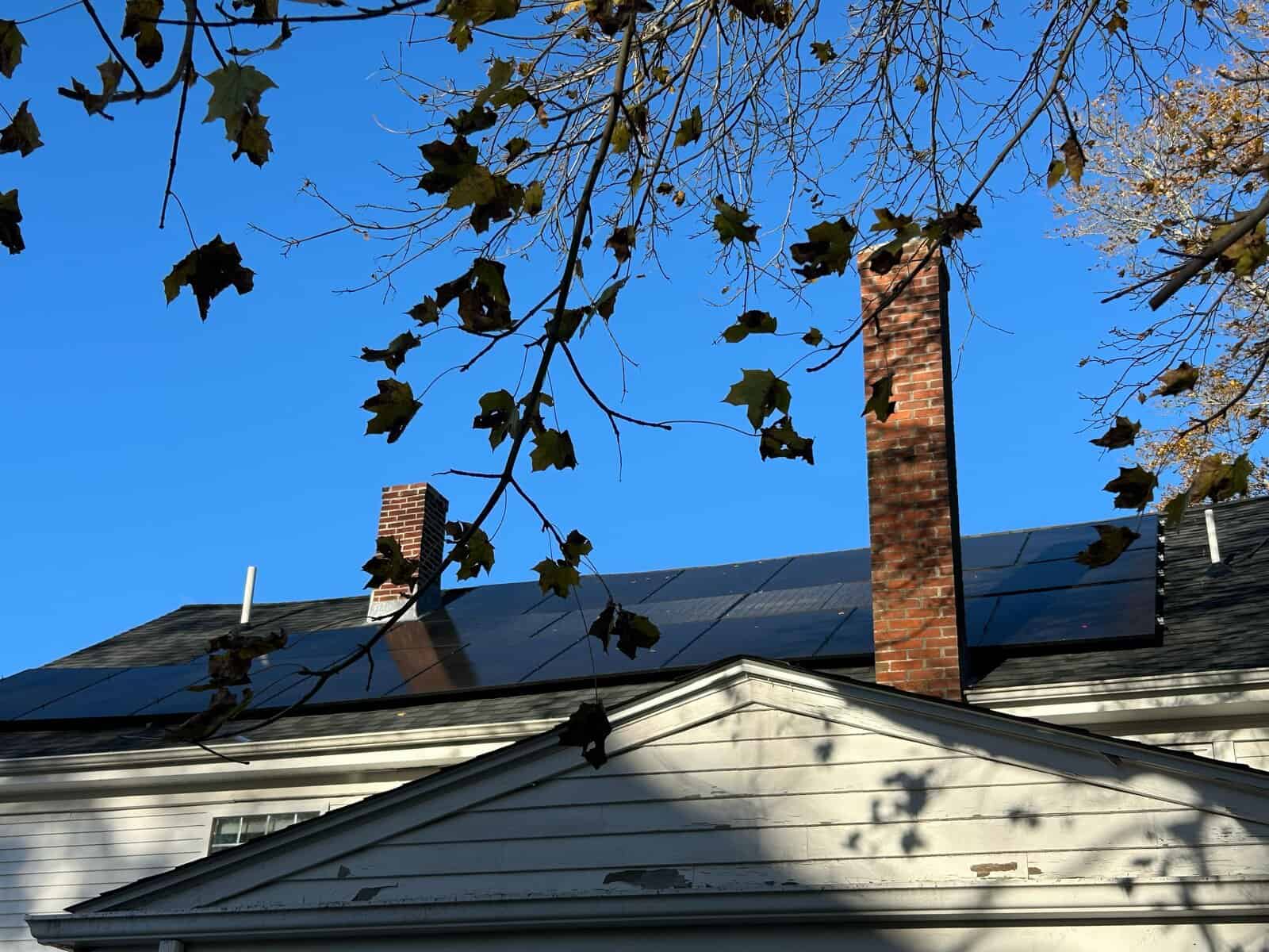 A close, upward-angled view of a home’s roof with a sleek solar array running along the ridge line, framed by bare autumn branches in the foreground. Brick chimneys rise through the roofline against a clear blue sky, with sunlight reflecting softly off the dark panels for a clean, seasonal New England feel.