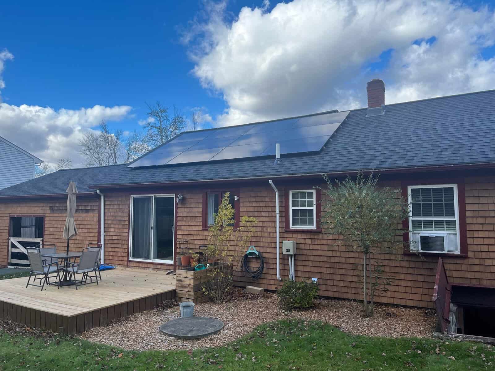 A rear-facing view of a single-story home with natural wood shingle siding, a small wooden deck and patio seating, and a clean, low-profile solar array mounted on the roof. The panels sit neatly above the deck area, framed by a bright blue sky with scattered clouds, light landscaping, and a quiet backyard setting.