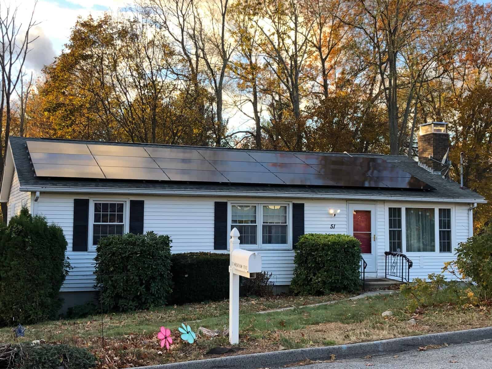 A front-facing view of a single-story white ranch-style home with black shutters, a red front door, and a full-width array of sleek black solar panels mounted along the roof, set against autumn trees with fall foliage and a mailbox in the foreground.