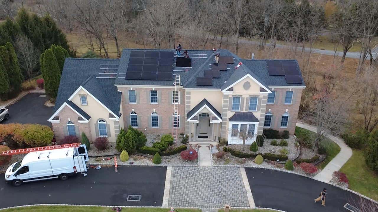 An aerial front-facing view of a large two-story brick home with multiple gables, showing solar panels in various stages of installation across the roof, a ladder positioned at the center, a white work van parked in the driveway, and installers working on the roof amid landscaped shrubs and a paved entryway, with leafless trees in the background.