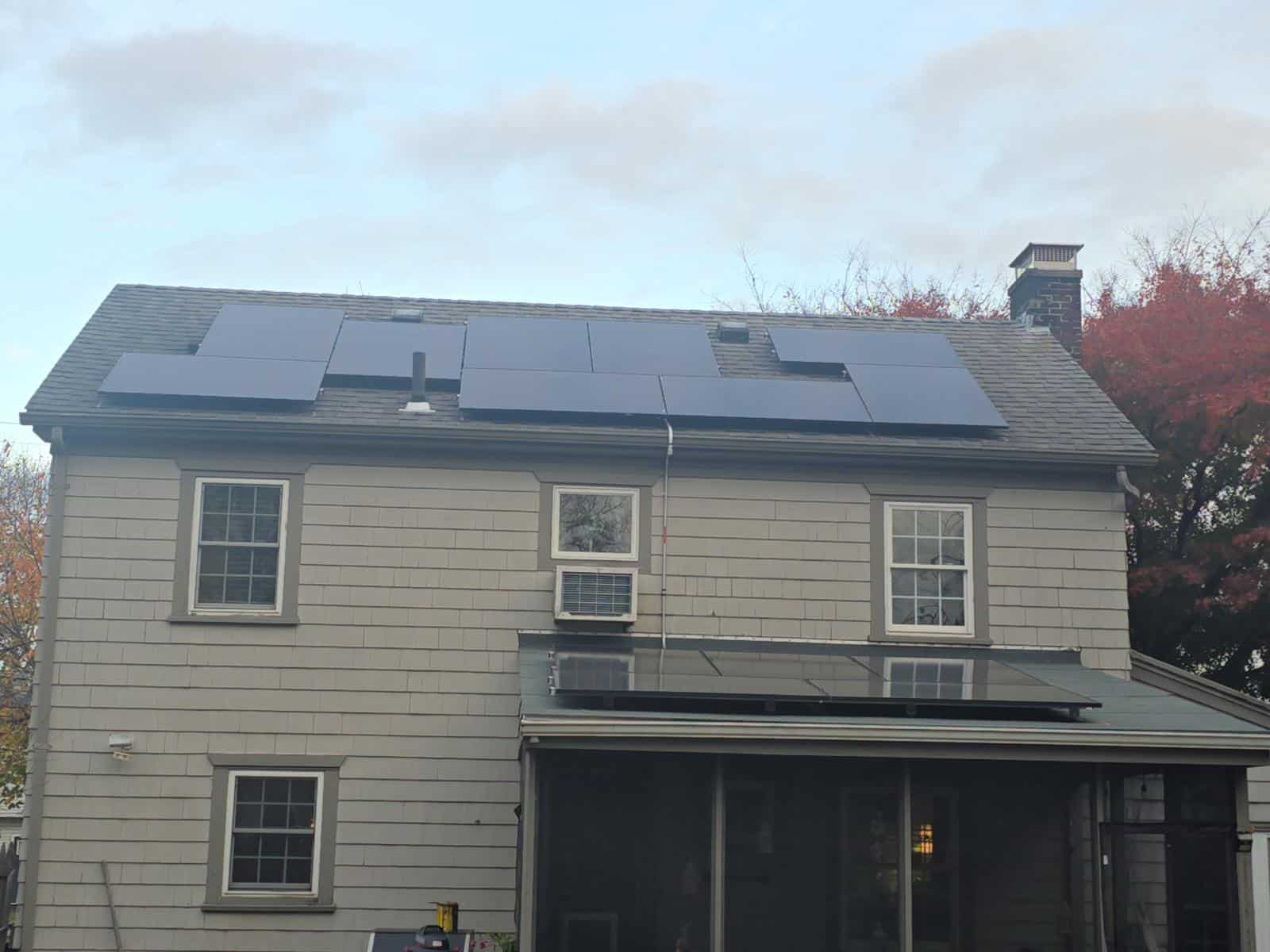 A rear view of a two-story gray shingle-sided home with a long roof-mounted solar array spanning most of the upper roof, a small porch roof with additional panels below, multiple white-trimmed windows, and a brick chimney on the right, set against fall foliage and an overcast sky.