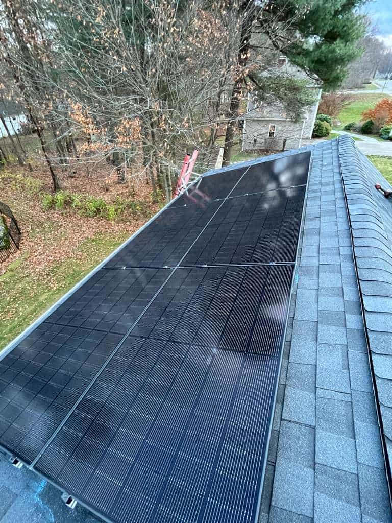 An angled rooftop view showing a low-profile black solar panel array mounted flush to gray asphalt shingles, with fall trees in the background and installation hardware visible along the roof edge.