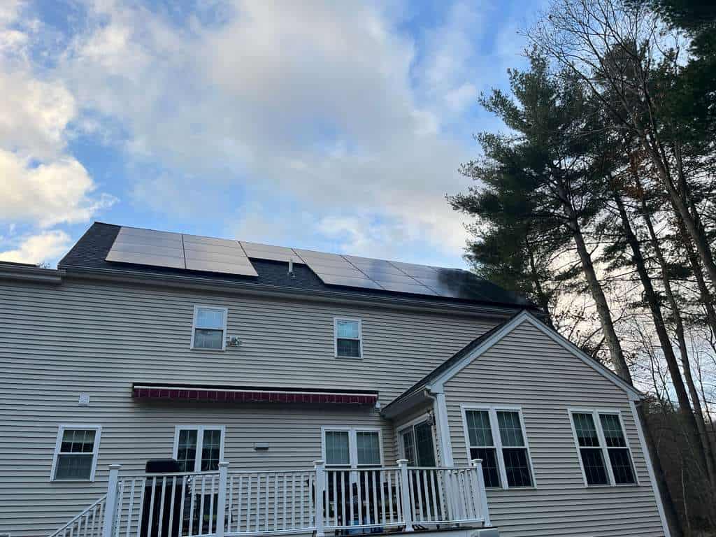 A rear view of a two-story beige-sided home with a long, low-profile black solar panel array mounted along the upper roofline, a white deck and railing below, and tall pine trees framing the house under a partly cloudy sky.