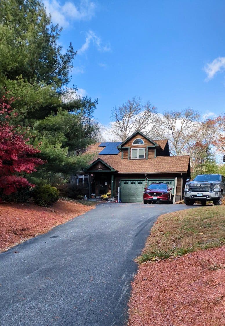 A front-angled view of a single-family home with brown shingle roofing and a small solar panel array mounted near the upper roofline, surrounded by mature trees with autumn foliage, a paved driveway, and two vehicles parked in front of a double garage under clear daylight.