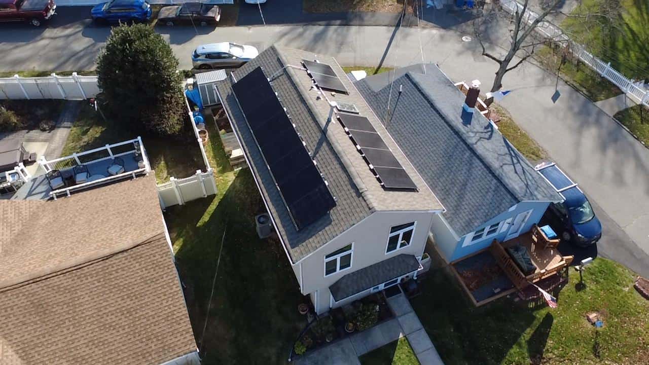 An aerial view of a residential neighborhood showing two adjacent homes with rooftop solar panel arrays installed along the main roof slopes, visible vents and chimneys, fenced yards, parked cars along the street, and clear midday sunlight casting sharp shadows.