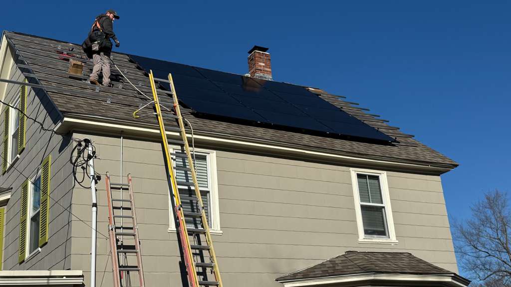 A rooftop installation scene showing a worker secured with safety gear standing on a shingled roof, positioning a large black solar panel array near a brick chimney, with ladders set against the house and tools visible during the install.