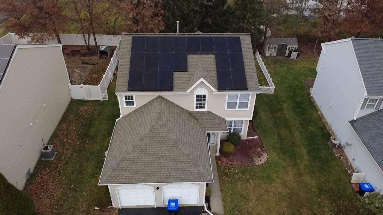 Aerial view of a two-story suburban home with a light-colored shingle roof, featuring a large black solar panel array arranged around a central roof section, with a front-facing garage below and neighboring houses, lawns, and trees visible on either side.