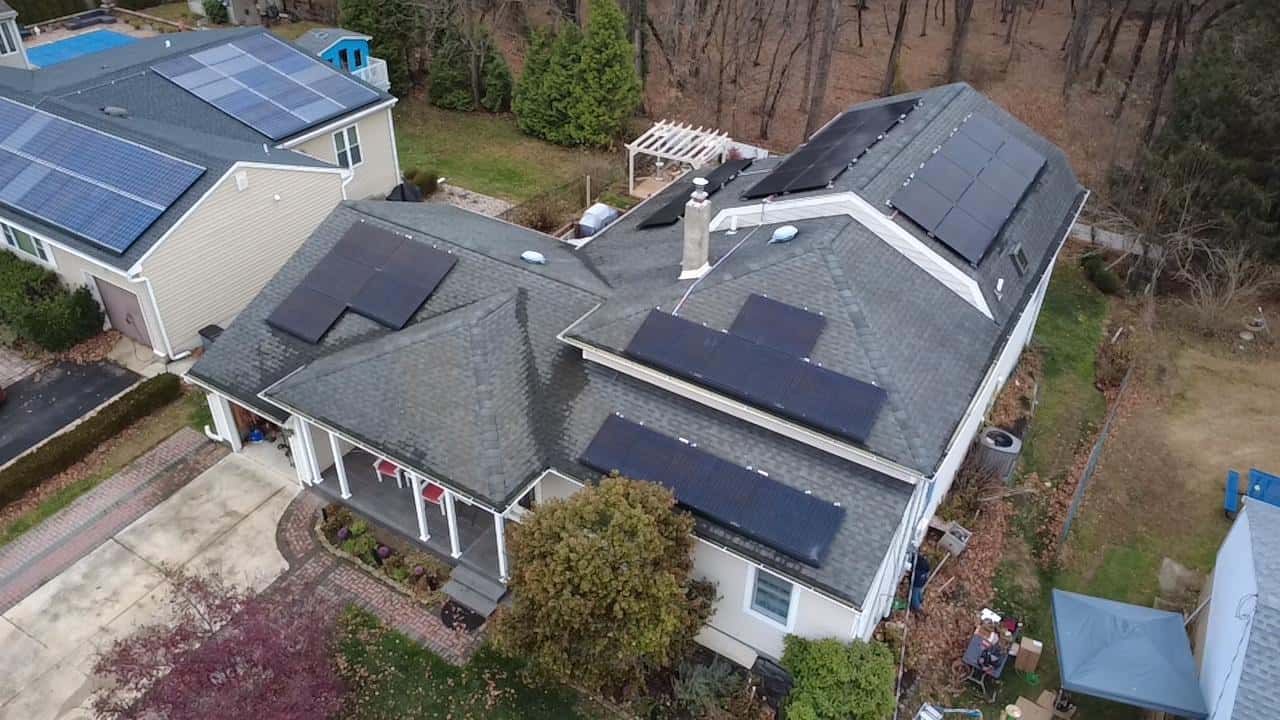 Aerial view of a large home with a complex roof layout, showing multiple black solar panel arrays installed across several roof sections, integrated around chimneys and dormers, with surrounding yard, neighboring homes, and trees visible.