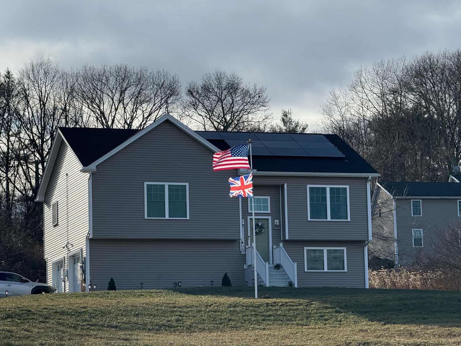 Front exterior view of a New England single-family home with gray vinyl siding and a black solar panel array mounted along the upper roofline, showcasing a clean, low-profile solar installation designed for year-round energy production in colder climates.