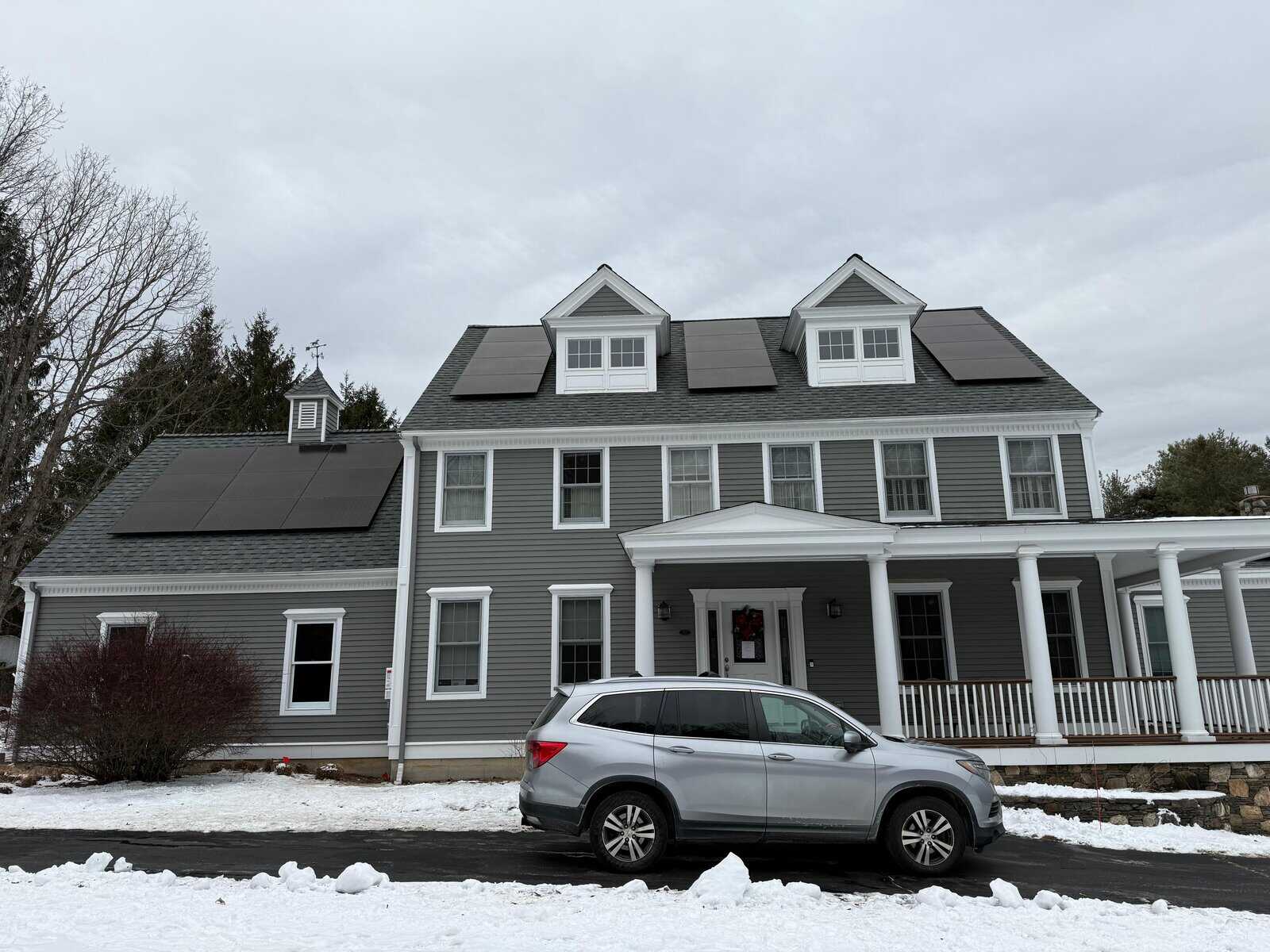 Front exterior view of a large New England–style home with a gray shingle roof and multiple black solar panel arrays installed across dormers and side roof sections, demonstrating a clean, balanced solar installation designed to perform reliably through winter conditions.