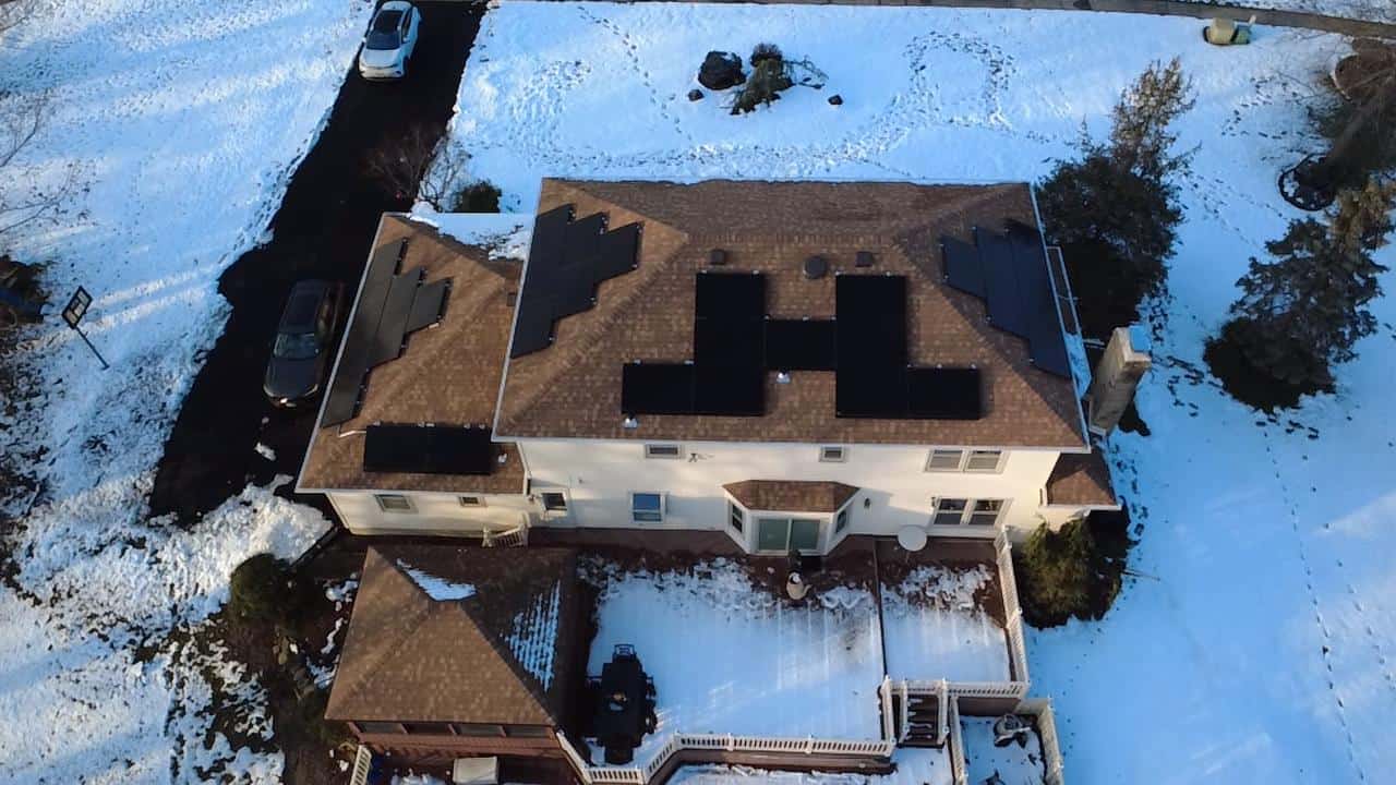Aerial view of a snow-covered residential property with multiple roof sections outfitted with black solar panels, showing a winter-ready solar installation integrated across a complex shingle roof layout.
