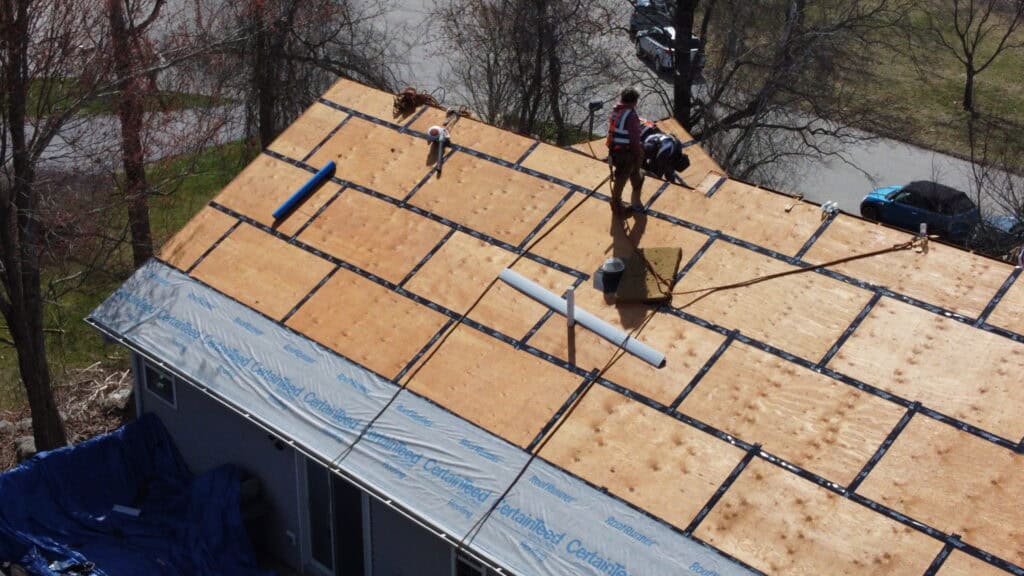 Worker installing roofing materials on a house, standing on a plywood roof deck with underlayment partially applied.